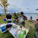 students at an outdoor table by the river examine water samples in a bag and tank