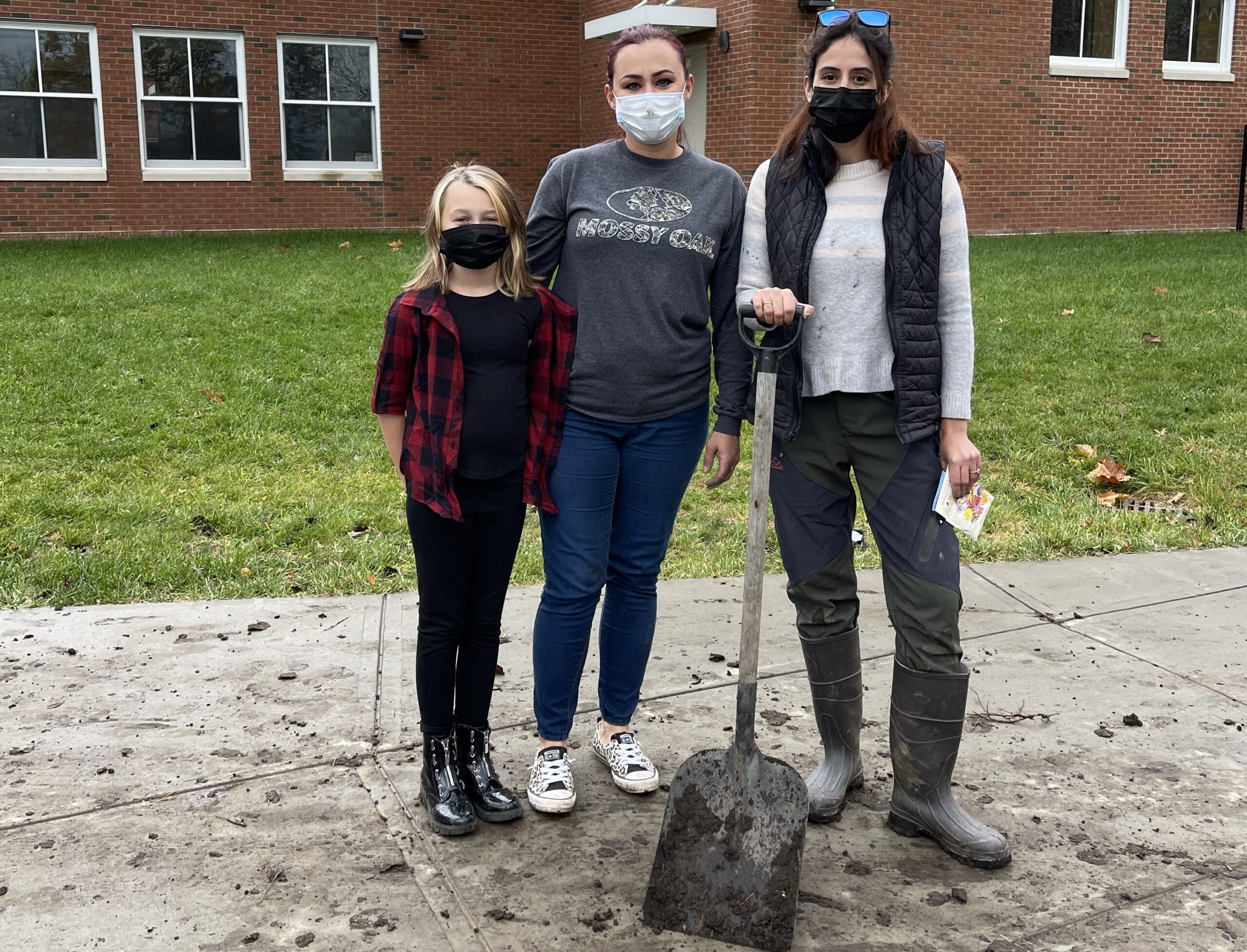 two adult females and a young girl stand in front of an elementary school and one adult is holding a large metal garden shovel
