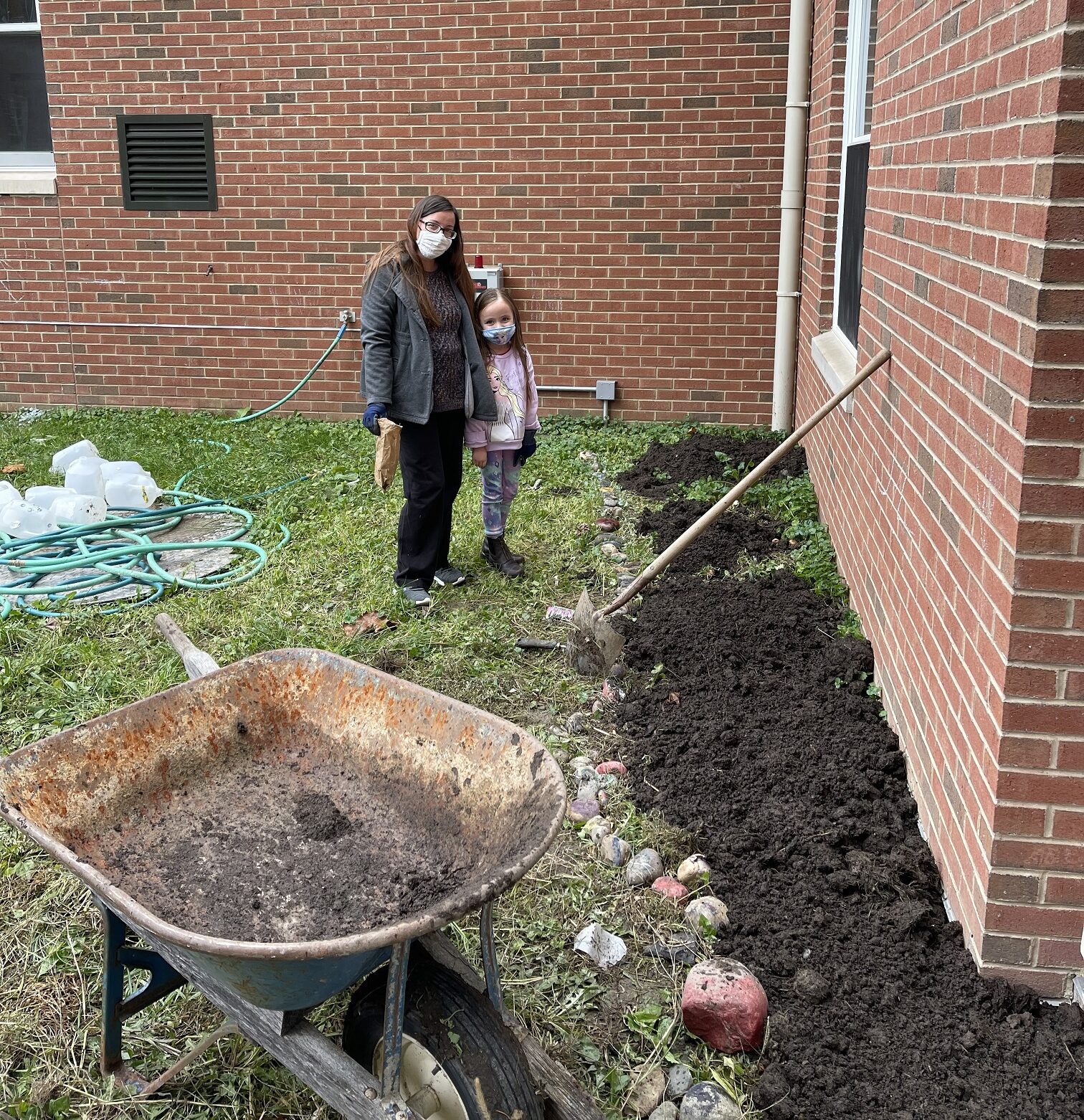 a woman and a young girl standing in the school courtyard with a shovel leaning on the wall and a wheelbarrow in the foreground