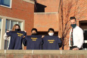 Mr. MacCormack stands with three elementary students by the school as they hold up Principals Award t-shirts