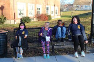 three elementary girls and one boy sit next to each other on an outdoor bench behind the school
