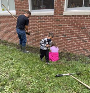 a man, young boy and young girl dig dirt in the school courtyard garden