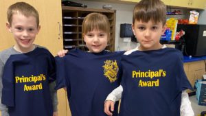 three caucasian children in a school office holding up their navy blue principals award tshirts