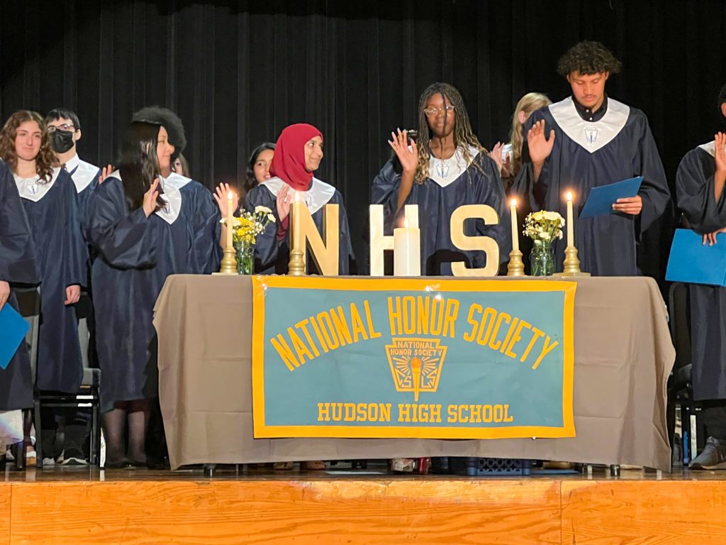 four high school students in dark blue gowns stand on auditorium stage behind a table with national honor society banner and they are holding up their right hands