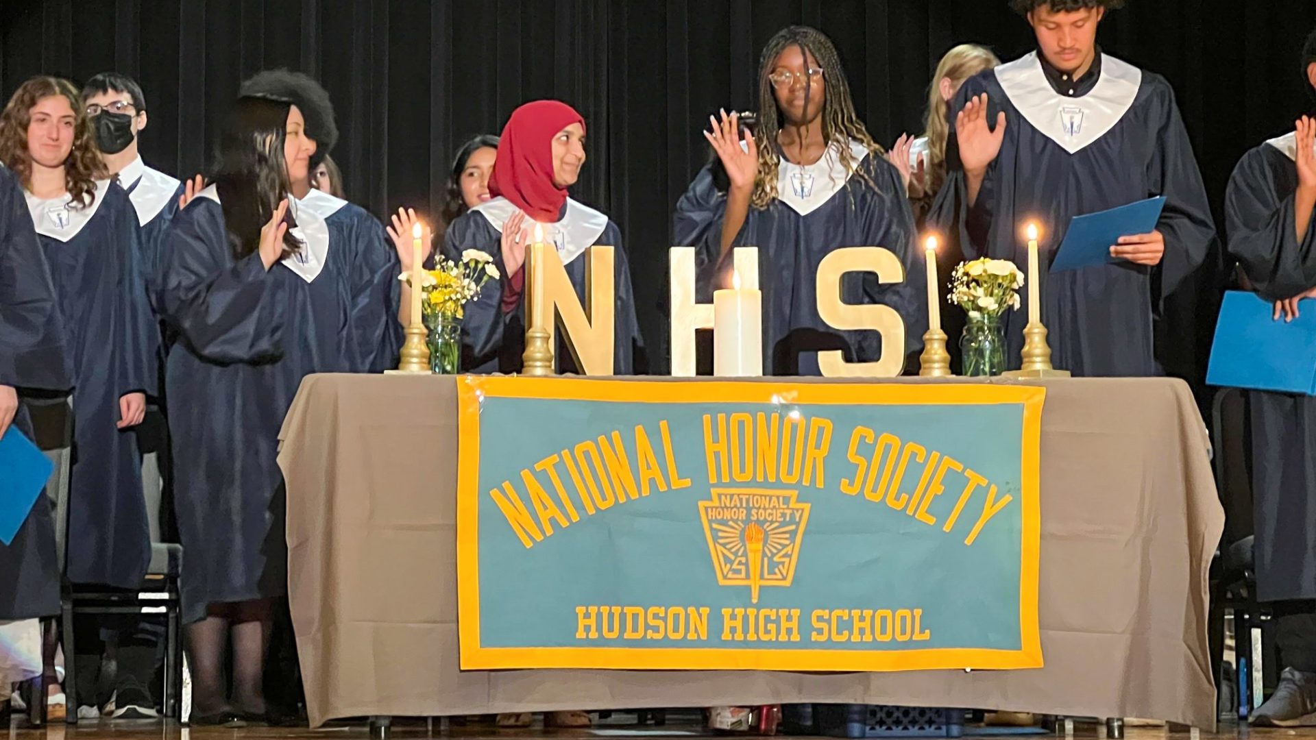 four high school students in dark blue gowns stand on auditorium stage behind a table with national honor society banner and they are holding up their right hands