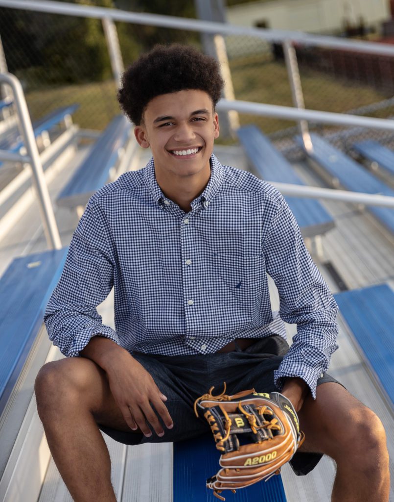 Isaiah Maines senior photo seated on outdoor bleachers and wearing a baseball glove on his left hand