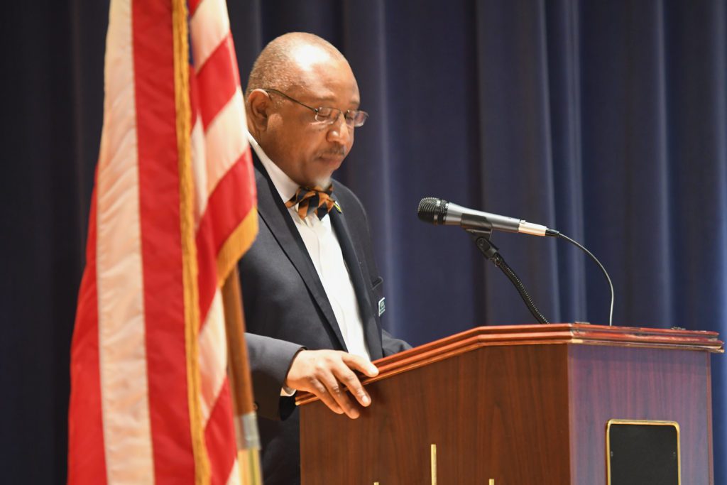 Dr. George Timmons speaks at a podium in front of blue stage curtains and an American Flag