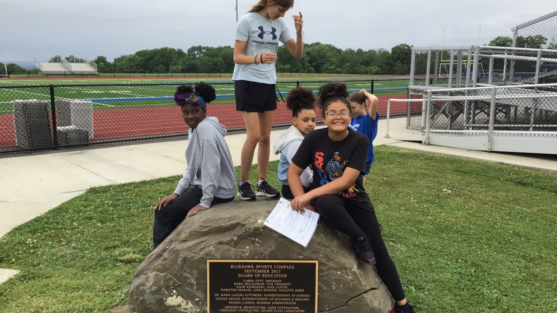 four junior high school girls standing and sitting on a boulder with a plaque situated in front of the track, field and bleachers