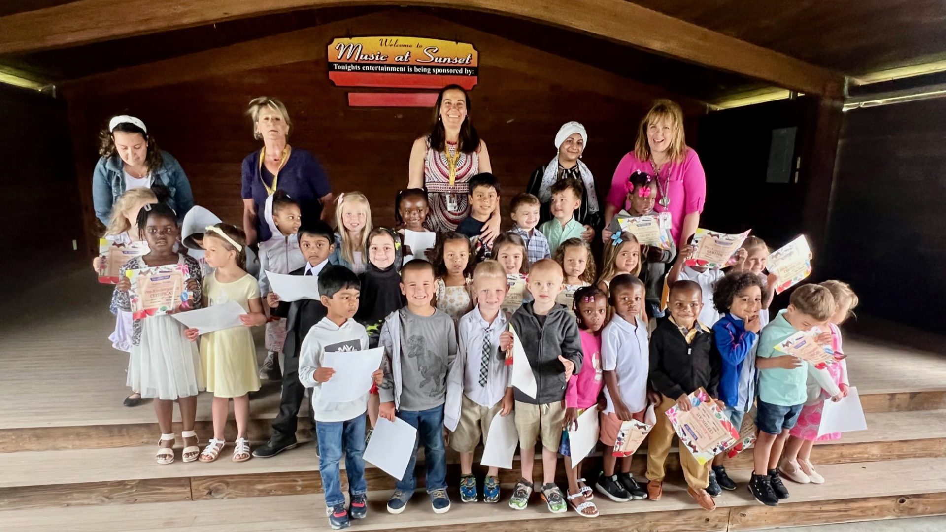 approximately 20 pre-kindergaten students holding certificates and pictures with five female adults standing behind them